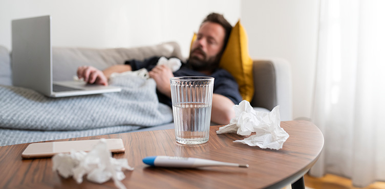 In the foreground, a coffee table with paper scraps, a fever thermometer, and a glass of water can be seen. In the background, which is blurry, a person is lying on a sofa with a computer.