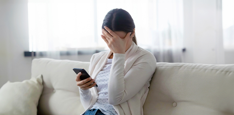 Kvinna som sitter i en soffa och tittar på sin mobil, ena handen döljer ansiktet.