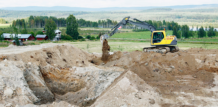 Excavator at a road construction site. A small farm and forest can be seen in the background.