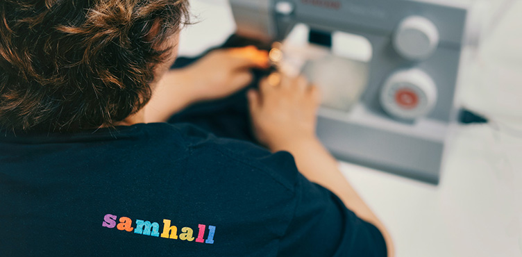 Detaljbild på person som syr på symaskin. På tröjan syns Samhalls logotyp.  