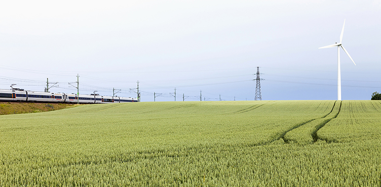 Ett fält med ett tåg, en kraftledning och ett vindkraftverk i bakgruden.