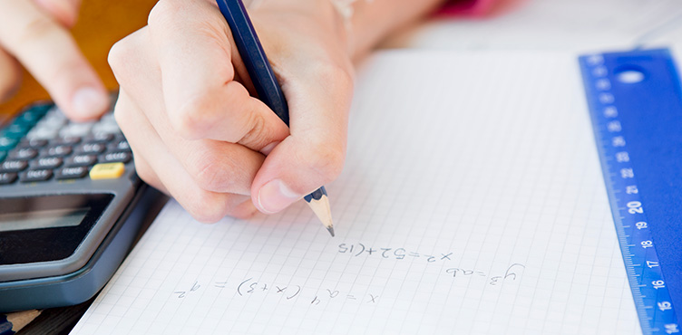 Detaljbild på hand som skriver ett matematiskt tal på rutat papper. Till vänster syns en miniräknare med ett finger på och till höger ligger en linjal.
