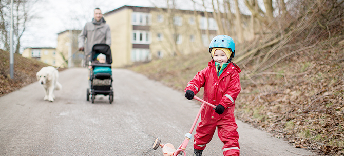 Barn med sparkcykel, pappa med barnvagn och hund på gata.