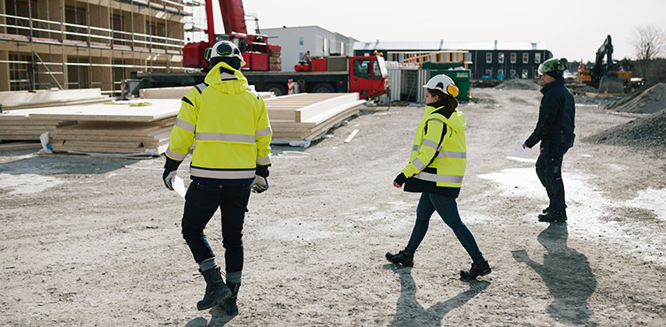 Three people can be seen at the construction site, two of them are wearing neon-colored jackets and helmets.