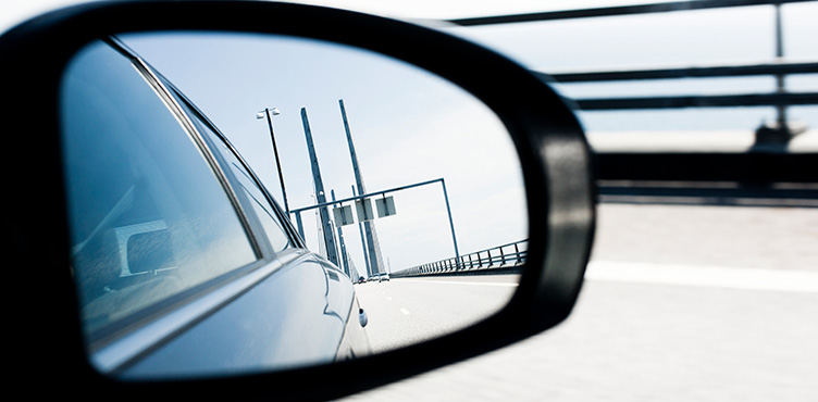 Närbild på backspegeln på en bil. I backspegeln ser man bropelare på Öresundsbron. 