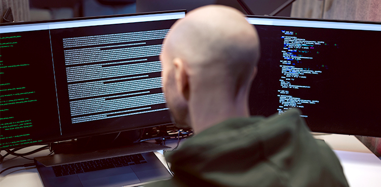 Man seen from behind at computer screens showing code.