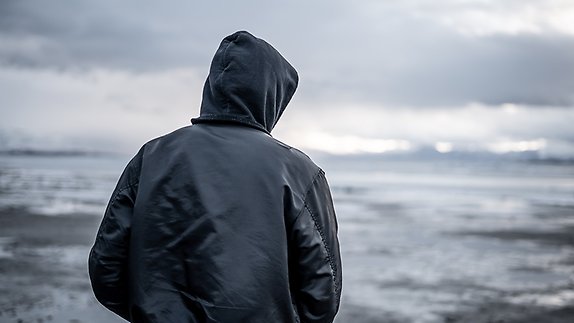 Person klädd i svart jacka med huva sedd bakifrån som tittar ut över grå himmel och vatten.