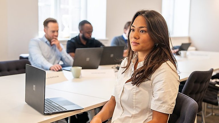 Ung person vid konferensbord som lyssnar på föreläsare. tre andra personer sitter på andra sidan bordet.