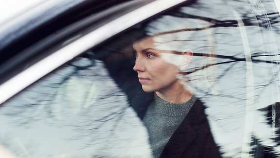 En kvinna sitter i en bil och tittar ut genom fönstret med ett eftertänksamt uttryck. Trädens grenar speglas i bilrutan.